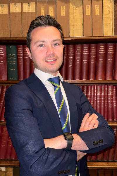Michael Duggan stands in front of a wall of bookshelves filled with red and brown hardcover volumes, wearing a navy suit, white shirt, and striped tie, with arms crossed and a confident smile.